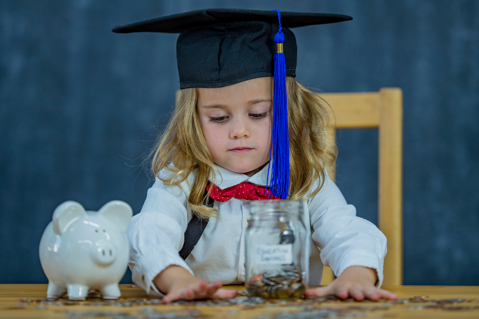 girl aged around 3 years wearing mortar board and bow tie, counting coins from a piggy bank